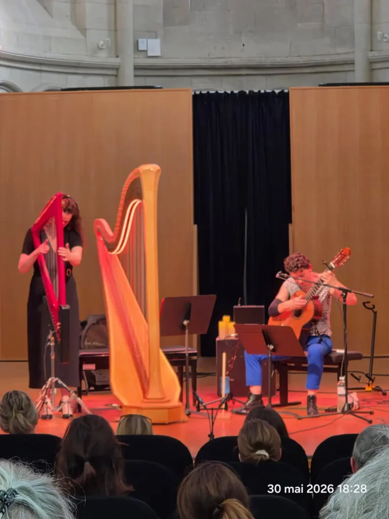 Delphine Baron à la guitare et Louise Ollivier à la harpe, sur scène lors d’un concert devant le public, dans une salle aux murs de pierre.