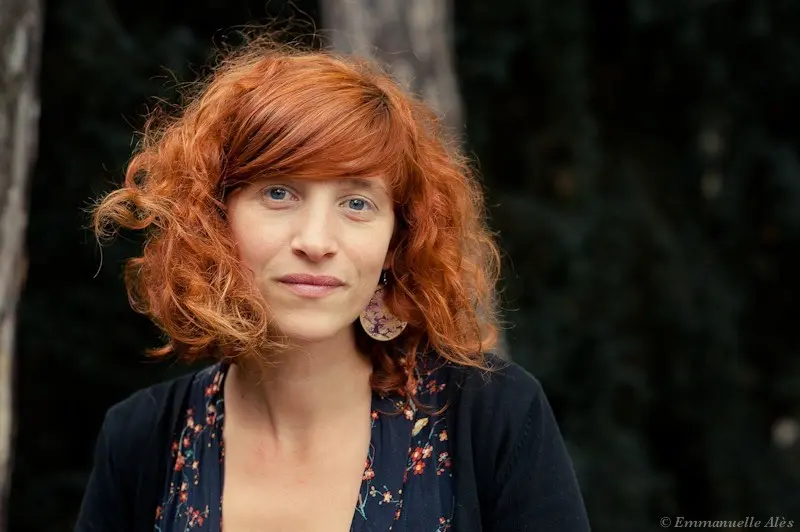 Portrait de Sandrine Marchetti, aux cheveux roux bouclés, portant une tenue sombre à motifs fleuris, devant un arrière-plan flou